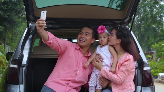 Happy family with their daughter sitting in the car and taking selfie photo with a mobile phone. Shot in 4k resolution