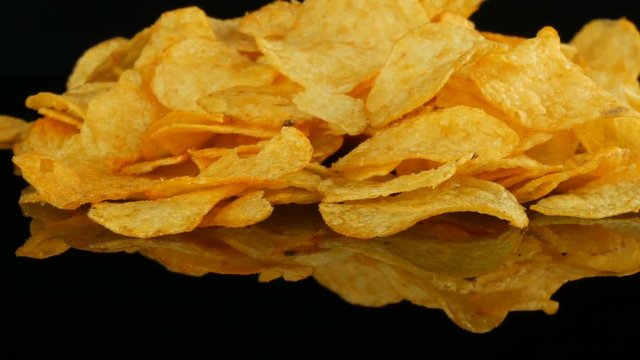 Potato chips lie on a mirror surface randomly scattered on a black background. Harmful food, fast food rotating.