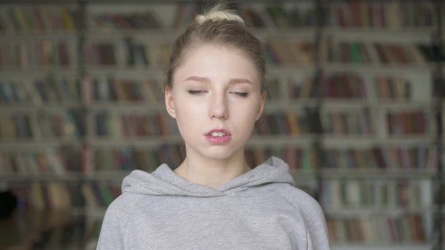 Portrait of tired beautiful female blonde student standing in library. Young girl green eyes in grey hoodie standing on the background of bookshelves in the library.