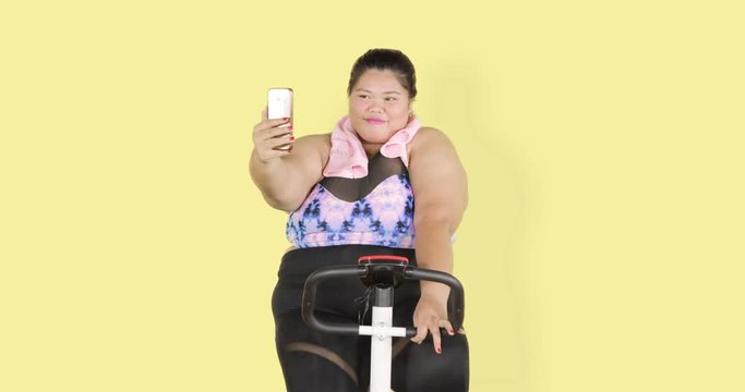 Overweight woman riding exercise bike while taking selfie picture with a mobile phone. Shot in 4k resolution
