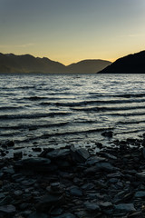 peaceful landscape during sunset with calm sky above the lake and mountain range, rocks in the foreground