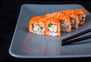 Sushi California Roll with crab meat, cucumber, masago, chopsticks on a gray plate, black background, side view