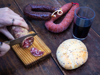 One person cutting an Iberian pork sausage on an old wooden board with an antique knife and rustic bread and a glass of red spanish wine