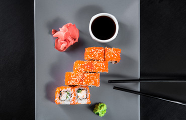 Sushi California Roll with crab meat, cucumber, masago, soy sauce, wasabi, chopsticks and ginger on a gray plate, black background, top view
