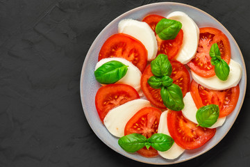 Italian caprese salad with sliced tomatoes, mozzarella cheese, basil, olive oil in a plate on dark concrete background
