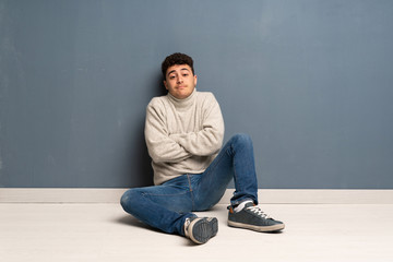 Young man sitting on the floor making doubts gesture while lifting the shoulders