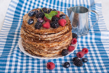 Pancakes on a platter with raspberries, blueberries and blackberries on a kitchen towel. A small rustic tin jug with milk. Lots of pancakes stacked. Thin pancakes with crispy crust. Carnival.