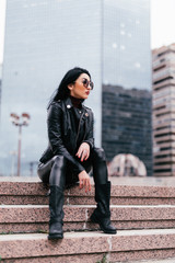 Fototapeta premium Casual style. Stylish woman in sunglasses sitting on the stone stairs. She dressed in leather jacket, skinny.