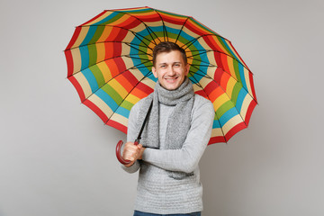 Attractive young man in gray sweater, scarf holding colorful umbrella isolated on grey background in studio. Healthy fashion lifestyle people sincere emotions, cold season concept. Mock up copy space.