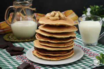 Pancakes with bananas on a plate against a dark background, behind is a jug and a glass of milk