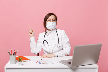 Female doctor sit at desk work on computer with medical document in face mask in hospital isolated on pastel pink wall background. Woman in medical gown glasses stethoscope Healthcare medicine concept