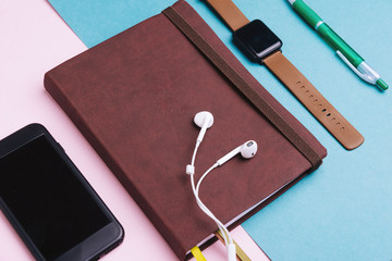 Flat lay photo of office desk with smartphone earphones, smartwatch and notebook on blue pink background