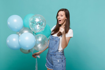 Funny young woman in denim clothes pointing index finger on camera, celebrating, hold colorful air balloons isolated on blue turquoise wall background. Birthday holiday party, people emotions concept.