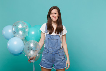 Concerned upset young woman in denim clothes biting lips, celebrating and holding colorful air balloons isolated on blue turquoise wall background. Birthday holiday party, people emotions concept.