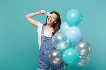 Funny young girl in denim clothes covering eye with credit bank card celebrating hold colorful air balloons isolated on blue turquoise wall background. Birthday holiday party, people emotions concept.