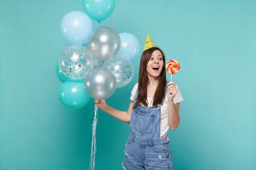 Surprised woman in birthday hat keeping mouth open, hold round lollipop, celebrating with colorful...
