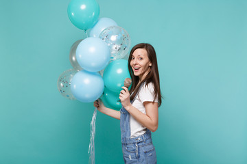 Side view of young woman in denim clothes sing song in microphone celebrating holding colorful air balloons isolated on blue turquoise wall background. Birthday holiday party, people emotions concept.