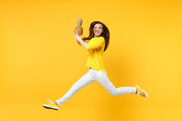 Smiling young woman in casual clothes jumping, hold fresh ripe pineapple fruit isolated on yellow orange wall background in studio. People vivid lifestyle, relax vacation concept. Mock up copy space.