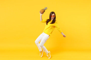 Fototapeta na wymiar Cheery young woman in heart glasses dancing, holding fresh ripe pineapple fruit isolated on yellow orange wall background in studio. People vivid lifestyle, relax vacation concept. Mock up copy space.