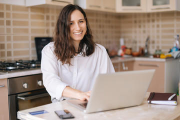 Smiling young woman using laptopand looking at camera in the kitchen at home