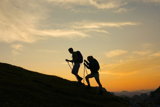 SILHOUETTE: Cheerful tourist couple are trekking up a grassy hill at sunset.