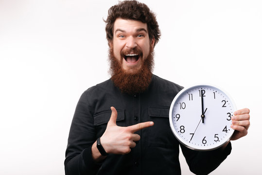 Handsome exicted bearded man pointing at white clock while smiling broadly at camera on isolated background