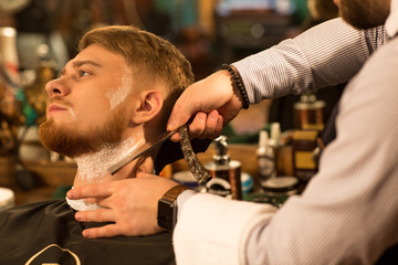 Close up of a young handsome bearded man looking away having his beard shaved by a professional barber with a razor service barbering profession occupation lifestyle hipster classic vintage retro well