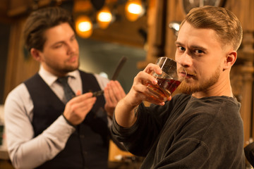 Young bearded handsome man looking to the camera fiercely drinking whiskey at the barbershop professional barber cleaning his razor on the background copyspace hipster service lifestyle masculine brut