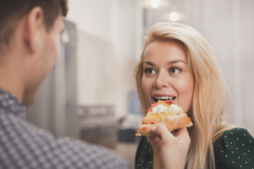 Beautiful young woman enjoying biting delicious pizza, her loving boyfriend offerng her. Man feeding pizza to his beautiful wife. Happy loving couple sharing pizza at home. Love, happiness concept