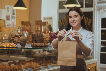 Beautiful cheerful female baker smiling, holding out paper shopping bag to the camera, working at her cafe. Lovely female confectioner selling delicious food at the bakery, copy space - obrazy, fototapety, plakaty