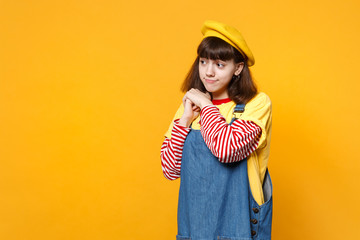 Portrait of confused pretty girl teenager in french beret, denim sundress looking aside isolated on yellow wall background in studio. People sincere emotions, lifestyle concept. Mock up copy space.