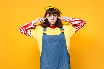 Portrait of upset girl teenager in french beret, denim sundress covering ears with fingers isolated...