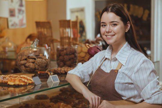 Cheerful young female baker smiling to the camera, working at the bakery store. Happy female waitress enjoying working at her cafe. Consumerism, friendly service, professions concept