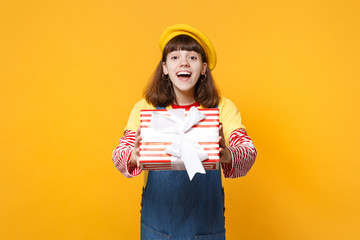 Cheerful cute girl teenager in french beret, denim sundress holding red striped present box with...