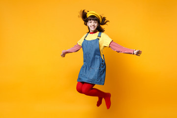 Portrait of smiling cute girl teenager in french beret and denim sundress jumping with flowing hair isolated on yellow wall background in studio. People emotions lifestyle concept. Mock up copy space.