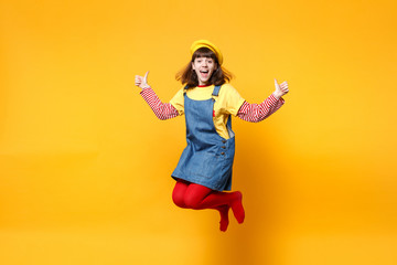 Fototapeta na wymiar Excited girl teenager in french beret, denim sundress showing thumbs up jumping and fooling around isolated on yellow wall background in studio. People emotions, lifestyle concept. Mock up copy space.