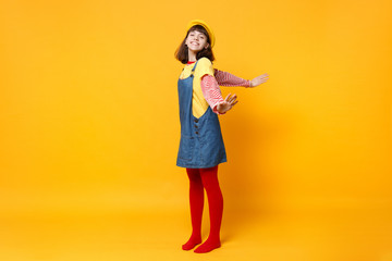 Full length portrait of charming girl teenager in french beret, denim sundress spreading hands isolated on yellow background in studio. People sincere emotions, lifestyle concept. Mock up copy space.