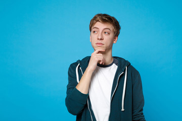 Portrait of pensive young man in casual clothes looking aside and put hand prop up on chin isolated on blue wall background in studio. People sincere emotions lifestyle concept. Mock up copy space.