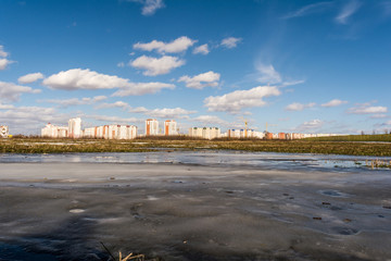 Fototapeta na wymiar view of the field with a frozen pond and the outskirts of the city, new city under construction, spring sunny day