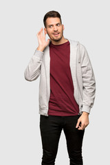 Man with sweatshirt listening to something by putting hand on the ear over grey background