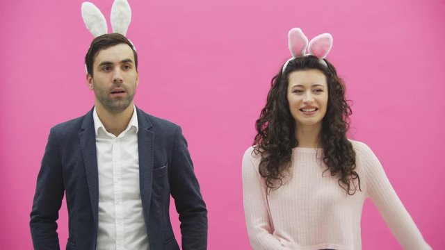 Young couple standing standing on pink background. During this time, they are dressed in rabble ears. Looking at each other smiles sincerely