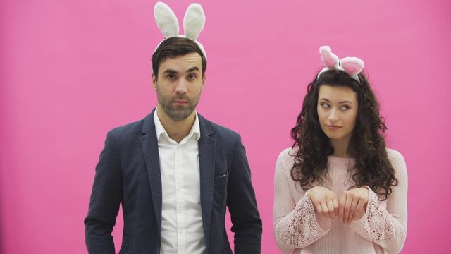 Young beautiful couple standing on a pink background. During this, they are dressed in rabble ears. Looking gently at each other, in love.