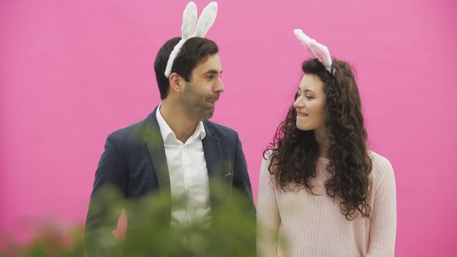 Young couple are beautiful on pink background. During this time, they are dressed in rabble ears. Looking at each other, behave like rabbits, reproducing movements of the mouth and teeth.