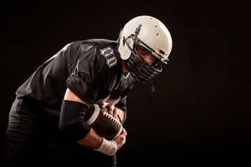 American football player in dark uniform with the ball is preparing to attack on a black background.