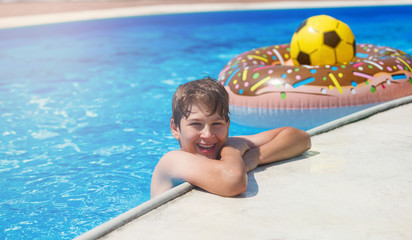 Happy cute little boy teenager lying on  inflatable donut ring with orange in swimming pool.  Active games on water, vacation, holidays concept. Chocolate donut. Cool fun summer holidays for children.