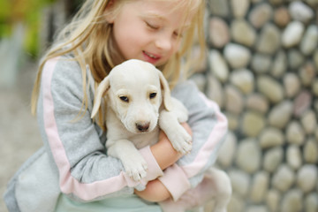 Cute little girl holding small white puppy outdoors. Kid playing with baby dog on summer day.