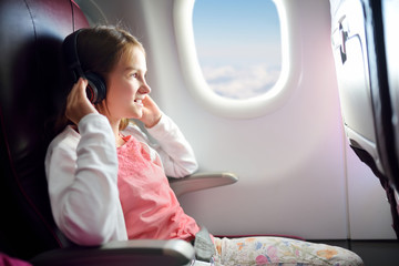 Adorable young girl traveling by an airplane. Child sitting by aircraft window and looking outside while listening to music. Traveling with kids.