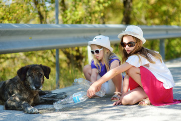 Thirsty black stray dog drinking water from the plastic bottle on hot summer day. Two kids giving cool water to thirsty dog.
