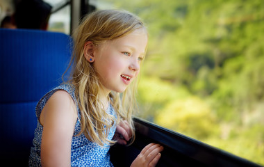 Cute young girl traveling by train on summer day. Child sitting by the window of railway wagon and looking outside.