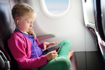 Adorable little girl traveling by an airplane. Child sitting by aircraft window and using a digital tablet during the flight. Traveling with kids.